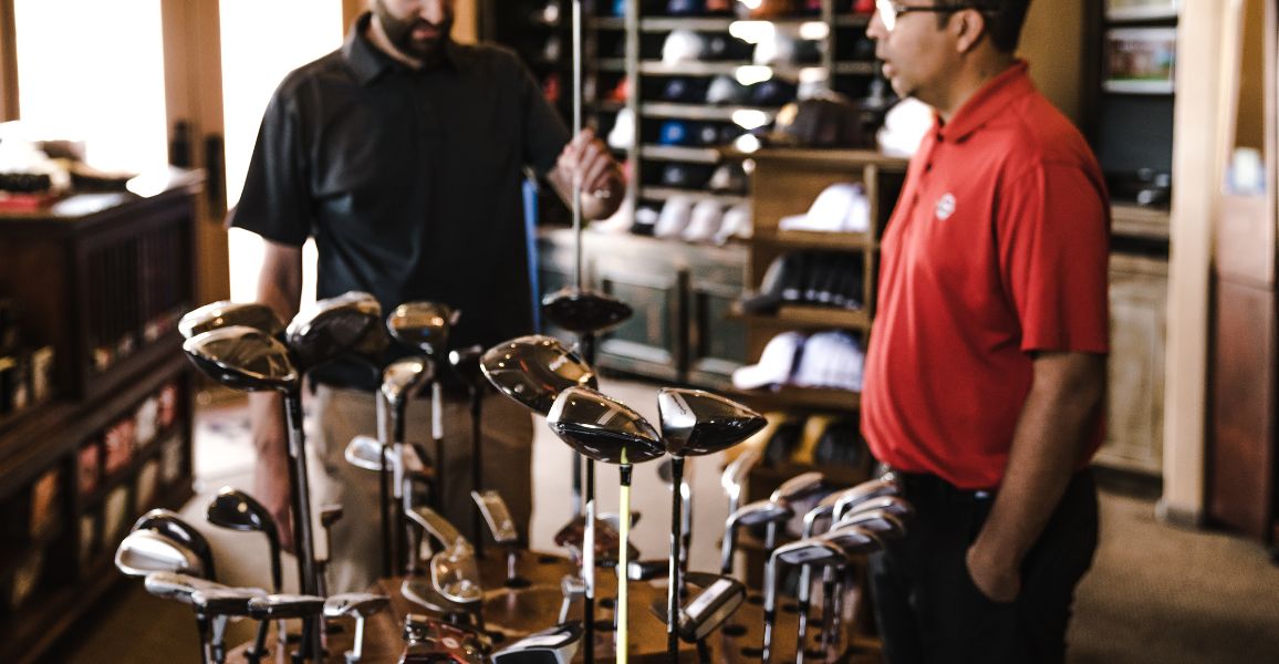 Golfer in a golfing shop looking at some clubs being helped by a man in red
