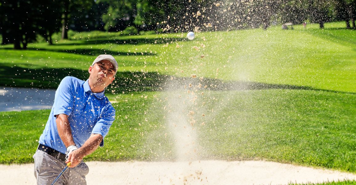 golfer dressed in blue hitting a bunker shot with sand flying through the air